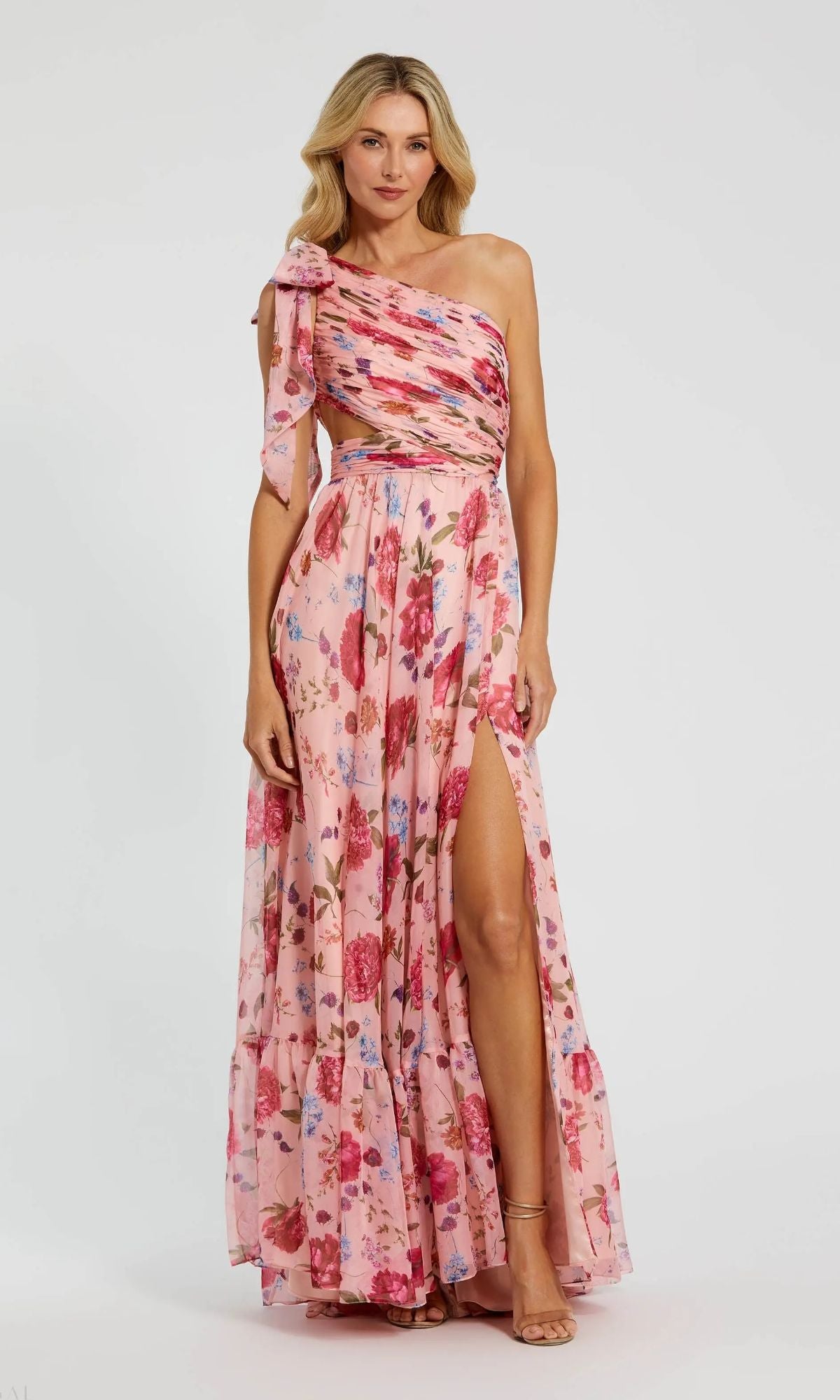 A woman wears the Mac Duggal 68553 long formal dress in blush multi, featuring a pink floral print, one-shoulder A-line design, and thigh-high slit, standing against a plain background.